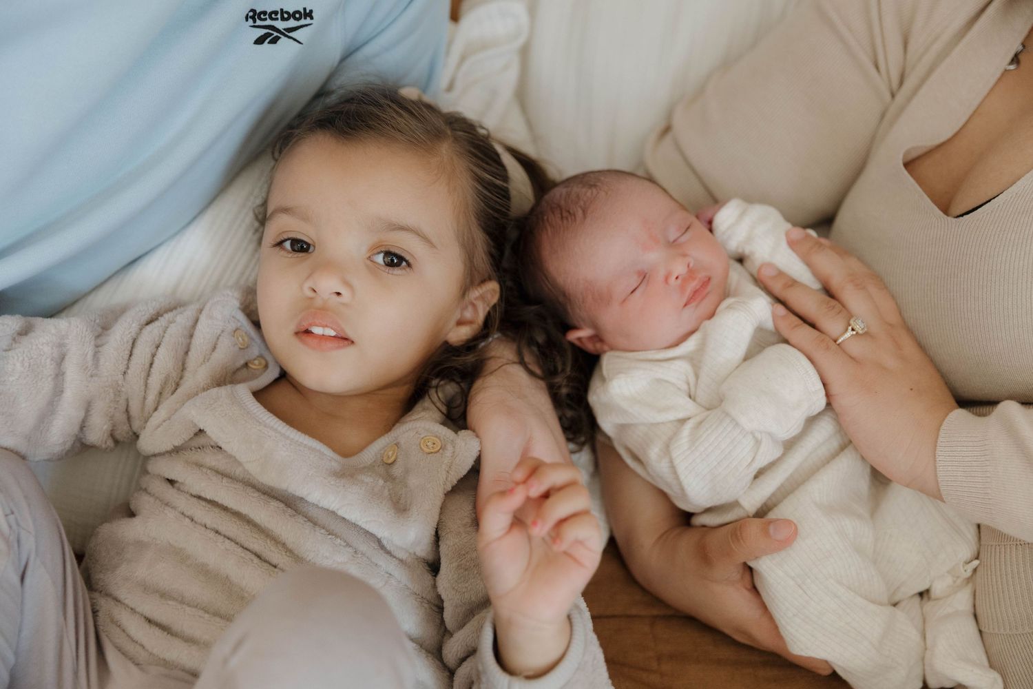 A toddler and newborn baby lying together on a couch in soft beige and white clothing.