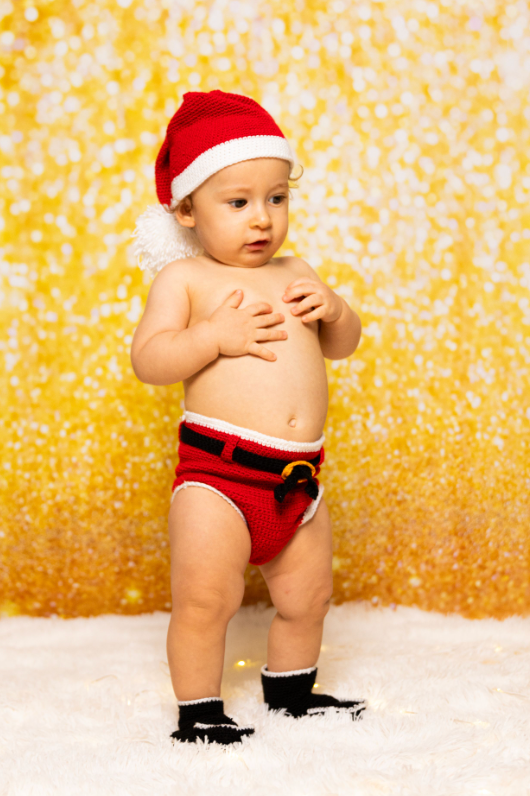Baby wearing red and black Santa outfit poses on snowy backdrop with golden bokeh lights.