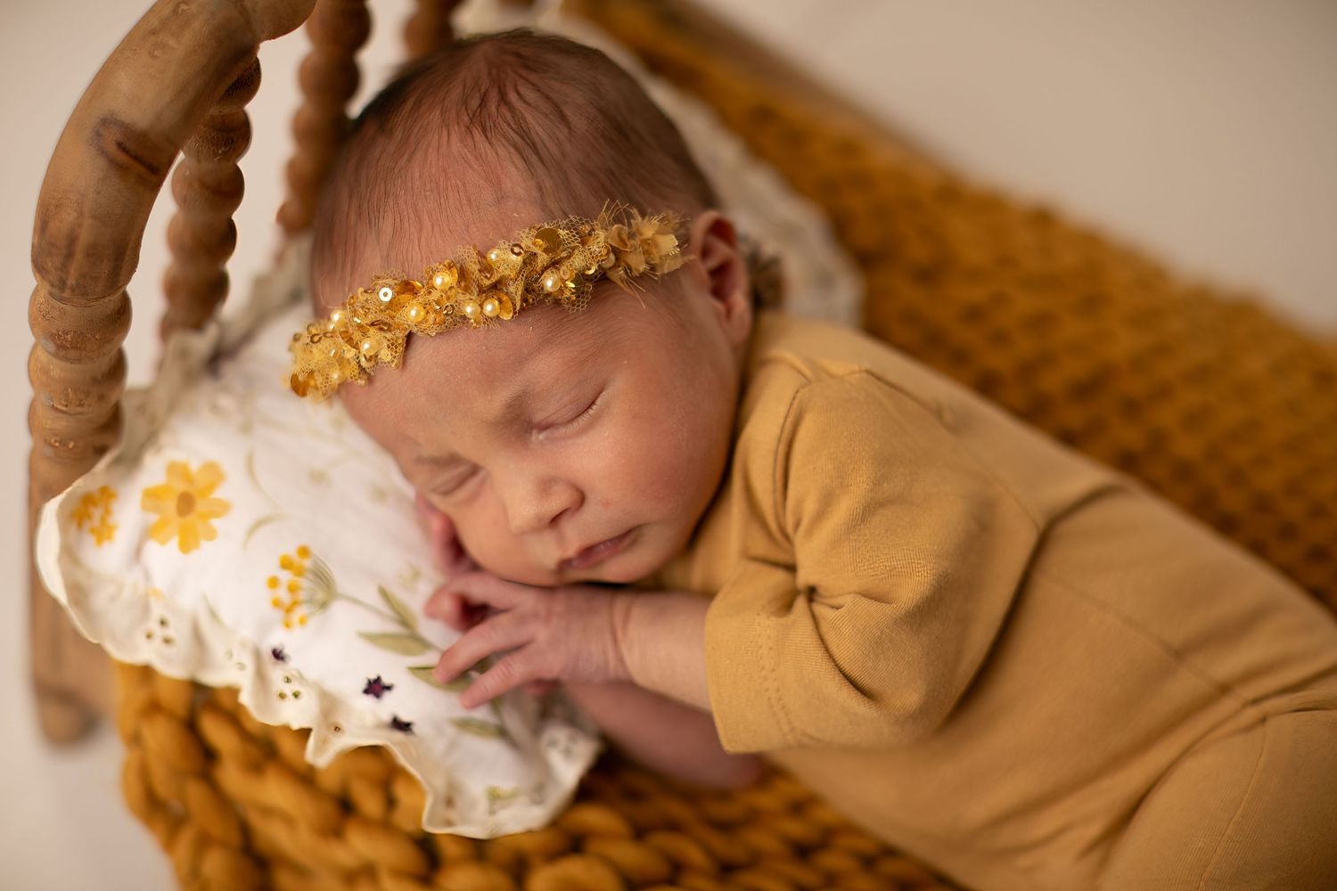 Sleeping newborn in mustard yellow outfit rests in woven basket with floral headband and white blanket.
