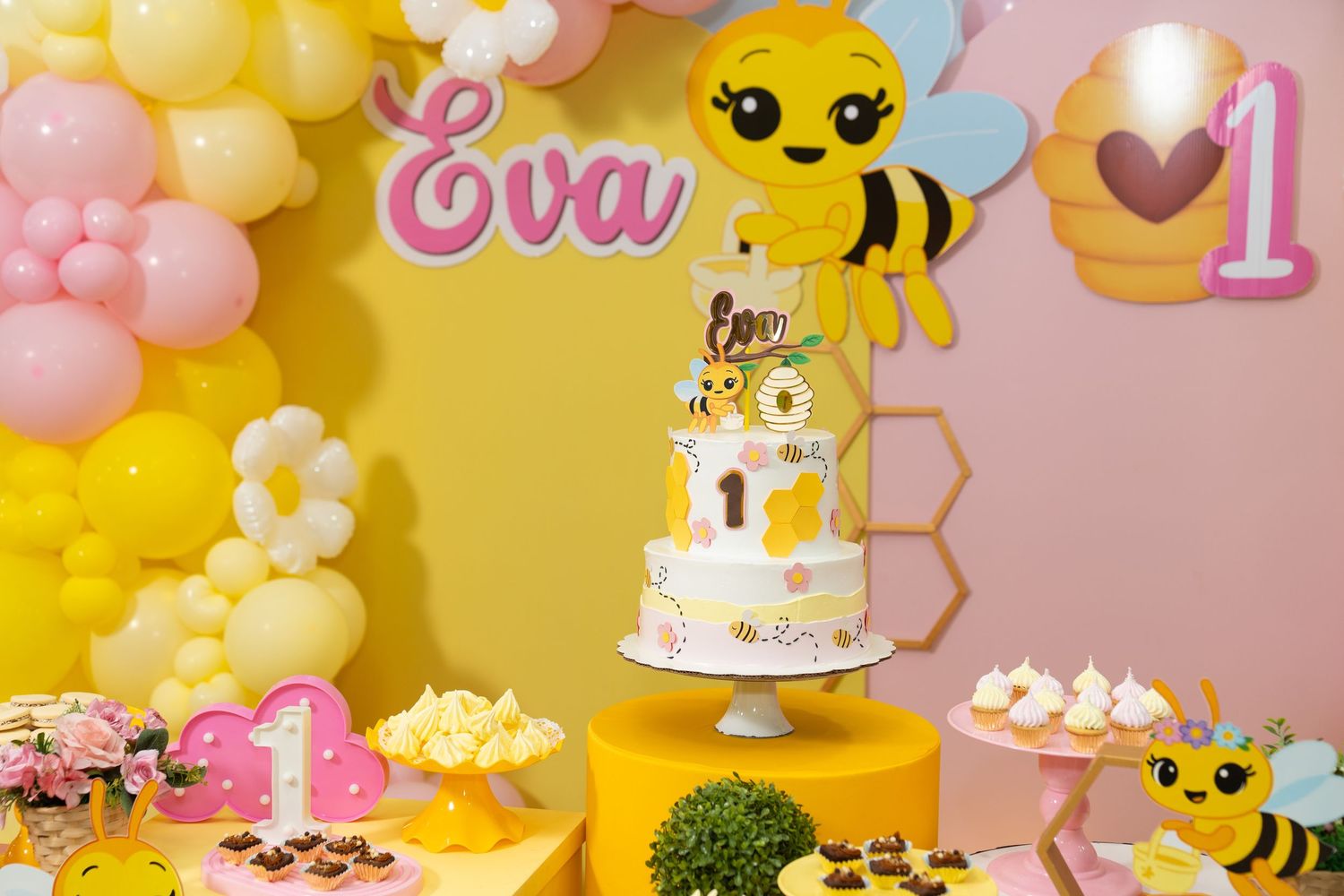 A pink and yellow themed birthday party setup with balloon decorations, a cake table, and bee-themed party elements.