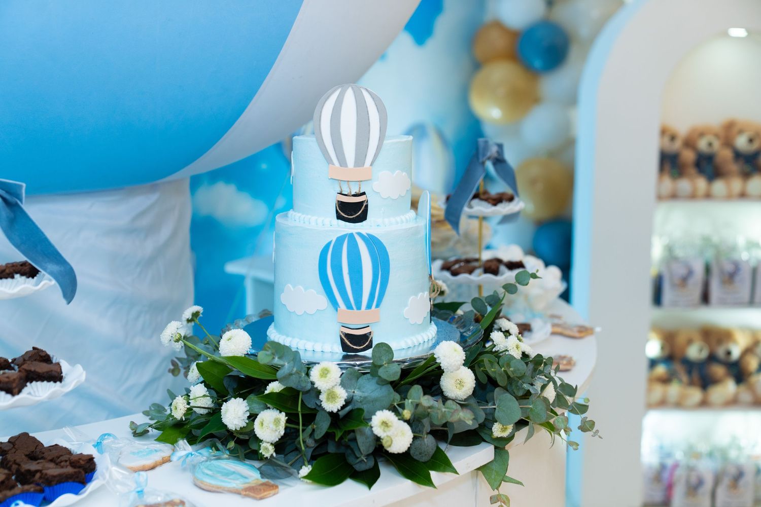 Blue and white hot air balloon themed cake with floral decorations on a dessert table at a party celebration.