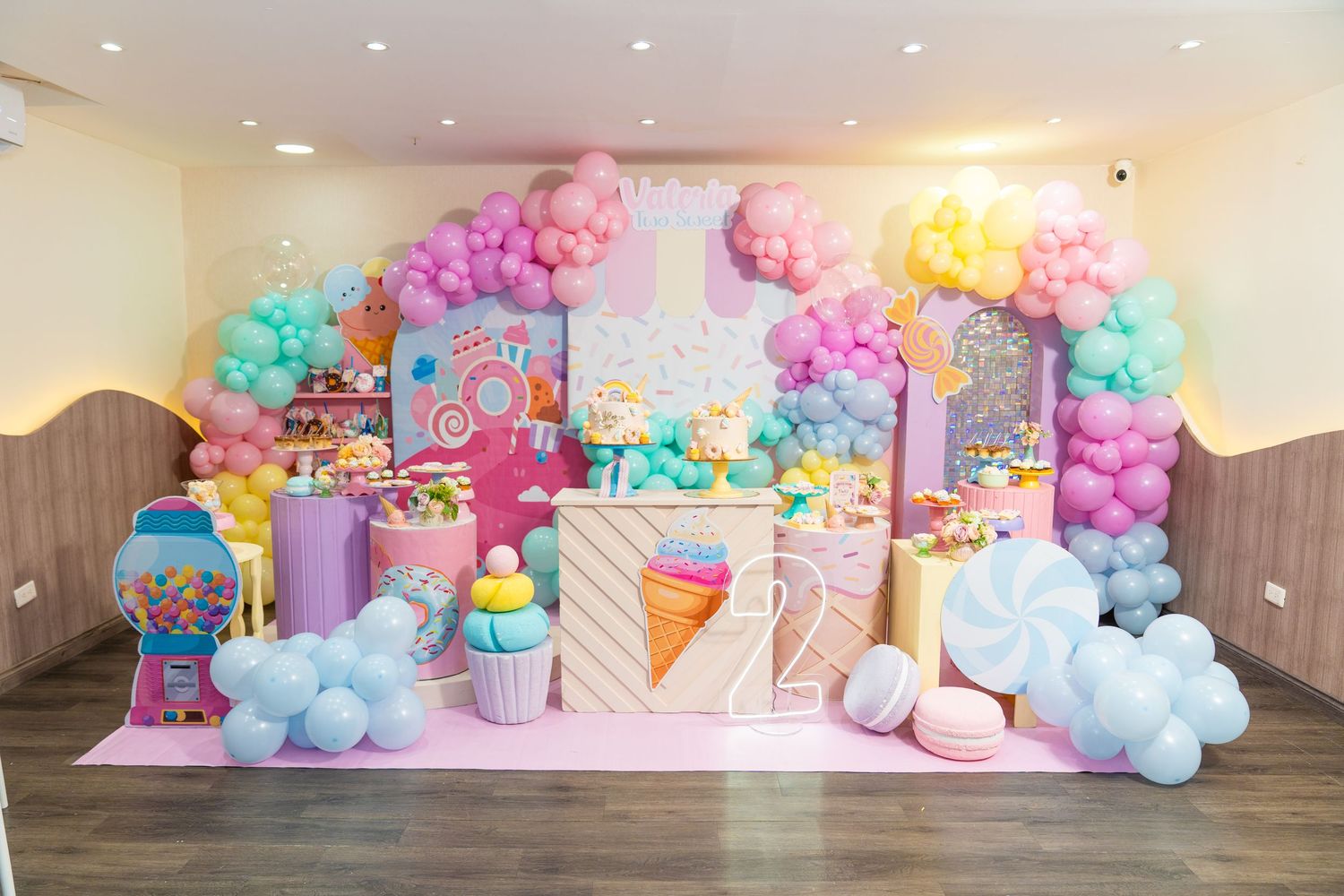 Colorful balloon arch and party decorations with pastel pink, blue, and yellow hues create a whimsical dessert-themed backdrop.