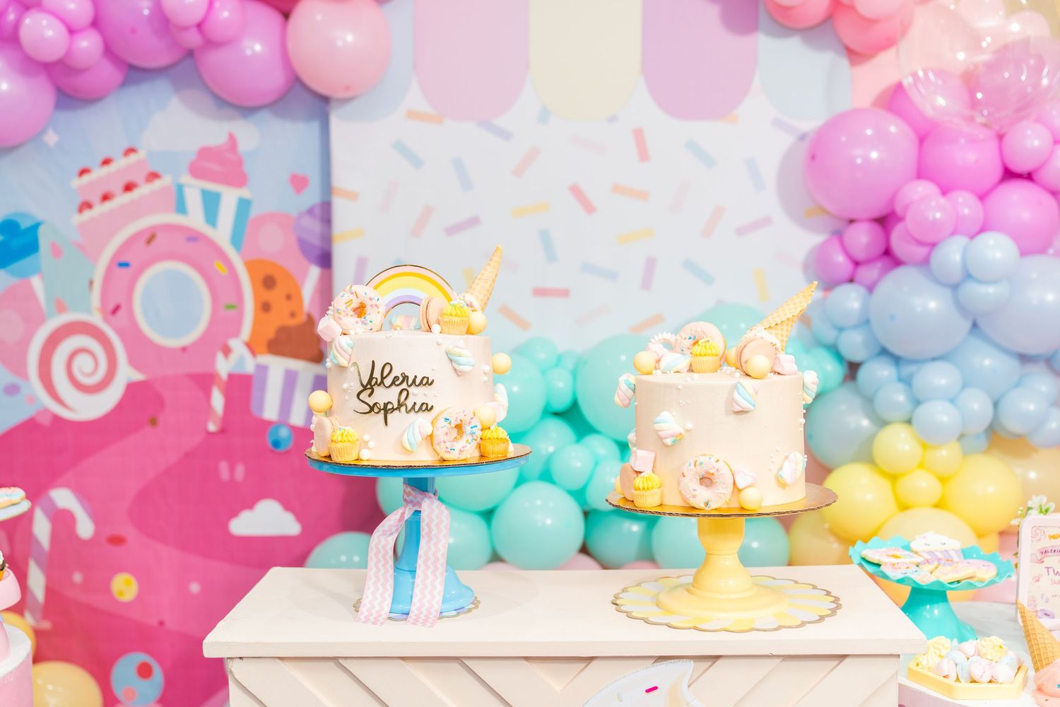 Pastel balloon arch decorates a dessert table with two elegant white and gold cakes on pink and yellow cake stands.