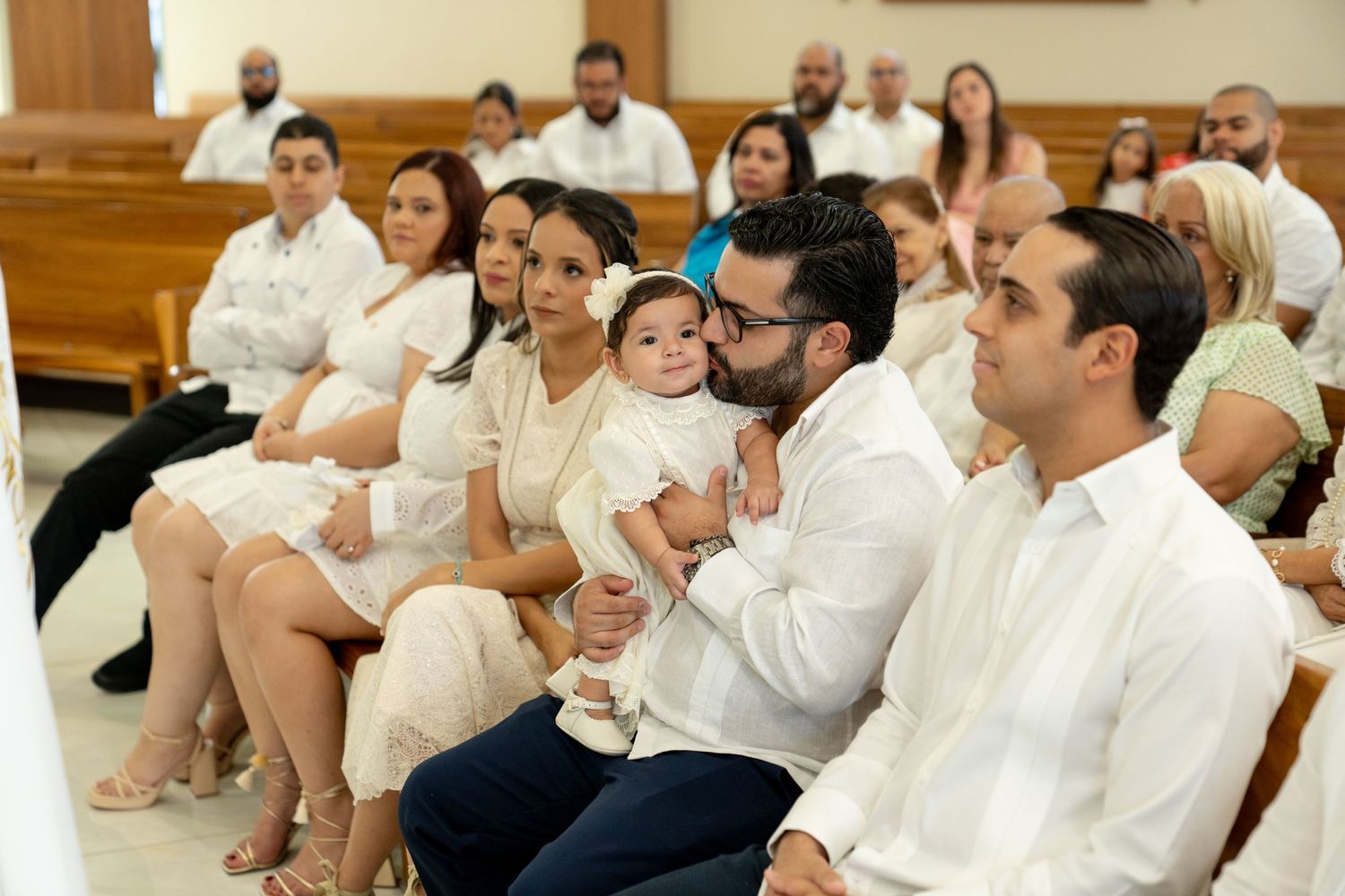 Attendees in white attire seated in wooden pews during a church ceremony.
