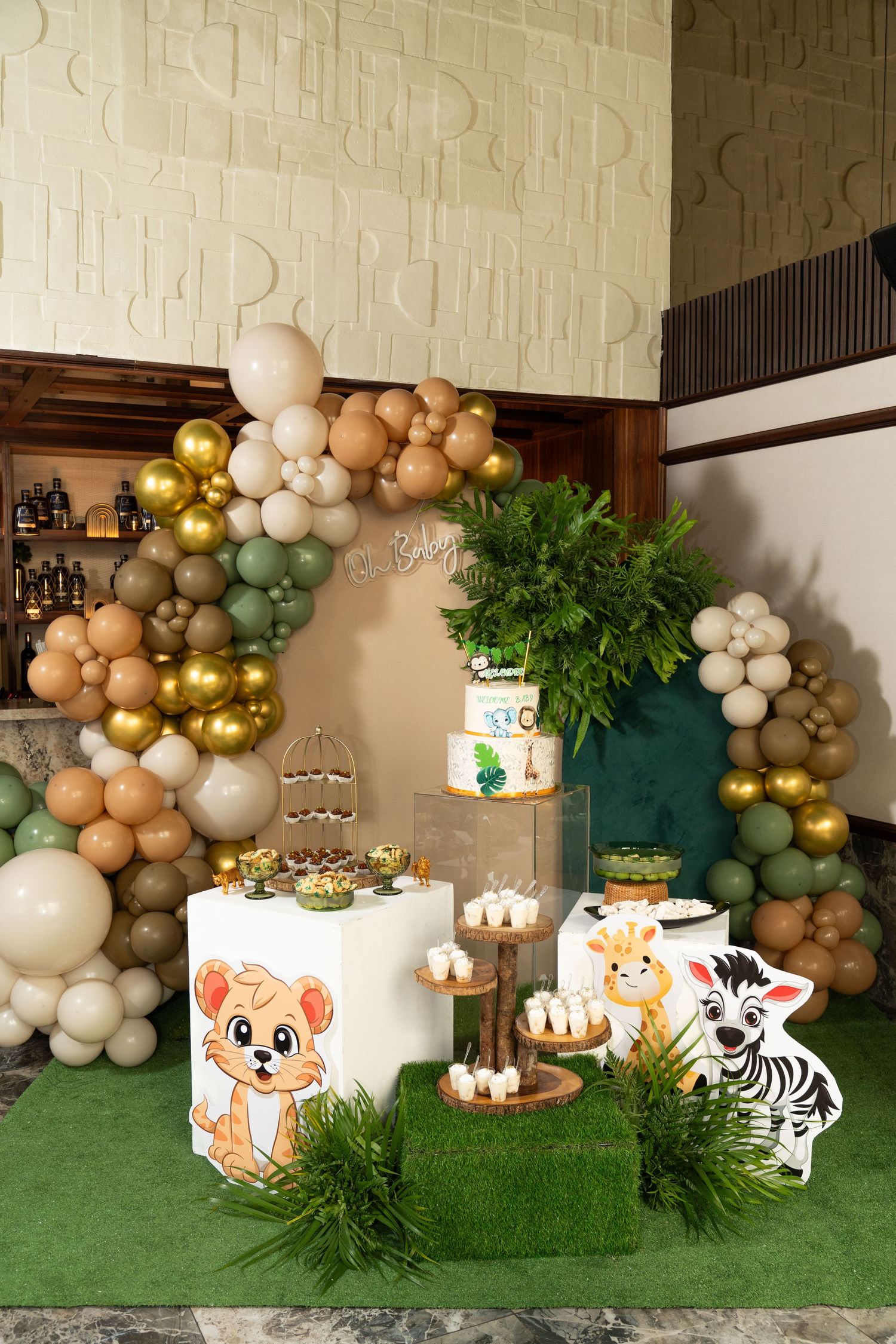 Safari themed party setup with balloon arch, zebra and fox cutouts, and green turf decorations against a textured wall.