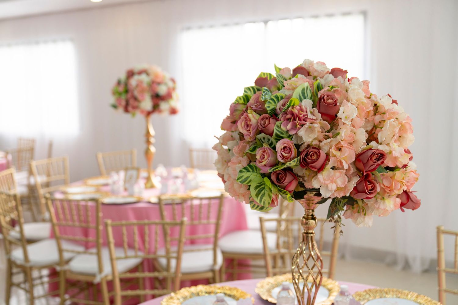 Elegant wedding reception table settings with pink floral centerpieces and gold candelabras in a bright white venue.