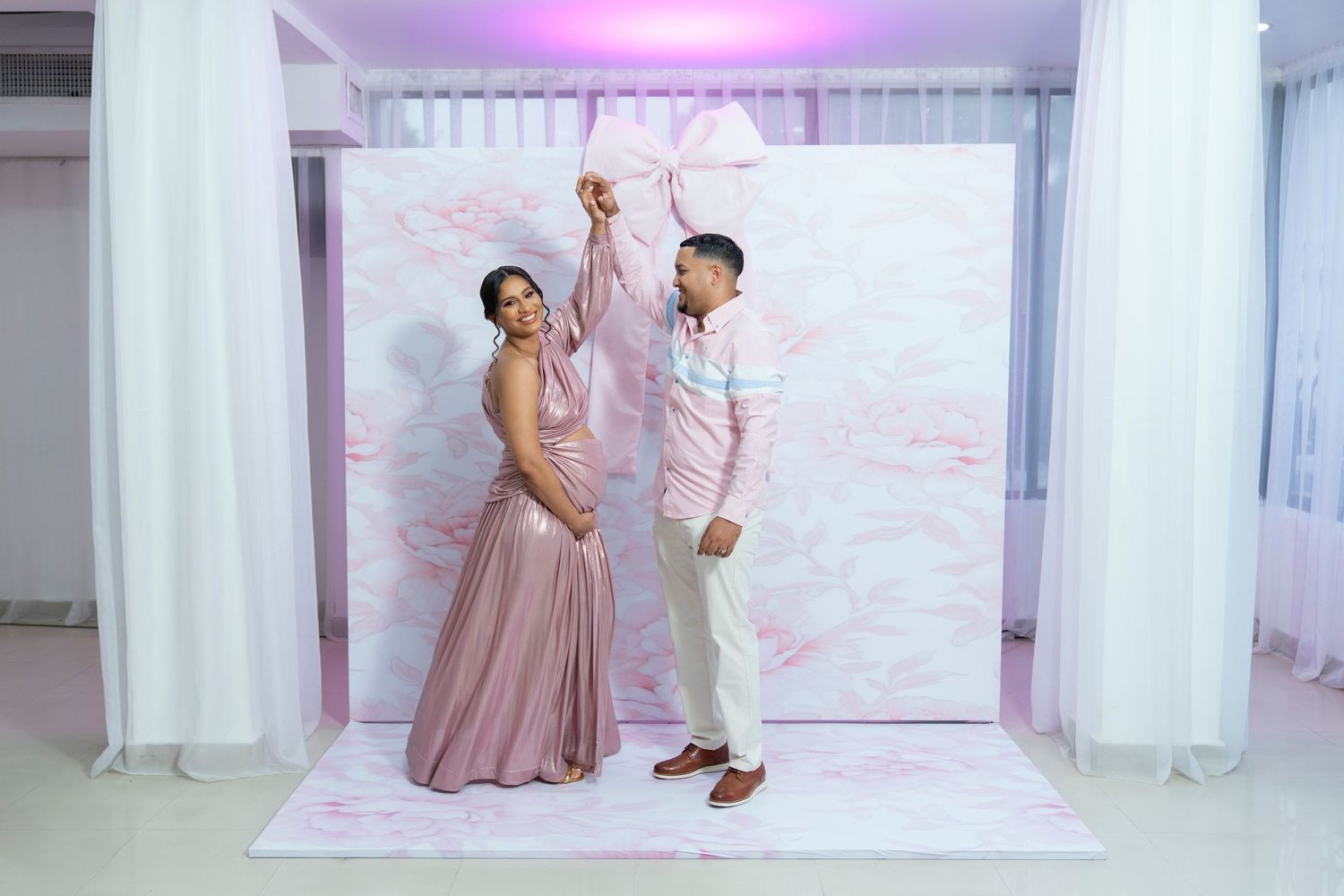 A couple dances together on a pink backdrop with white curtains and purple lighting during a special celebration.