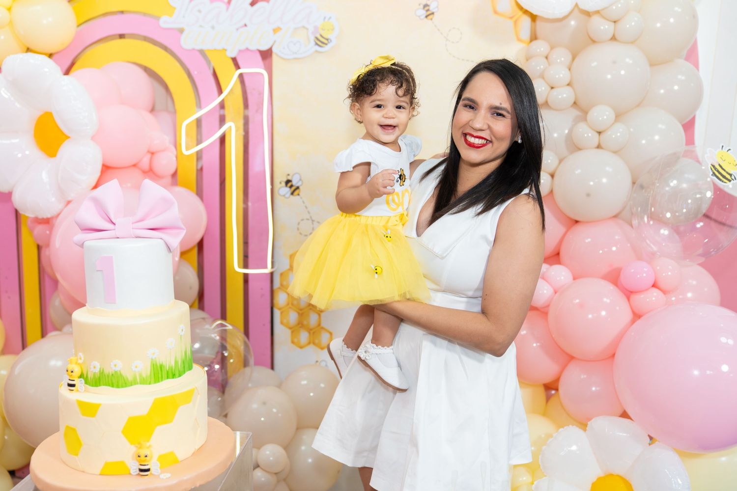 A festive birthday party setup with pink and yellow balloons, white decorations, and a birthday cake display.