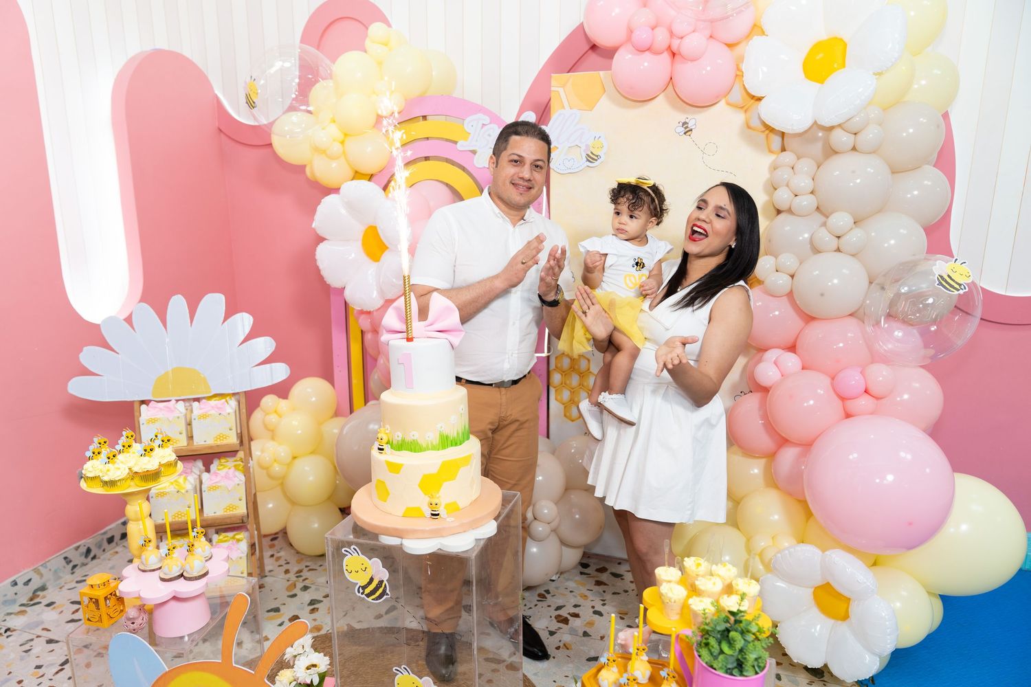 Festive party setup with pink and yellow balloon arch, daisy decorations, and dessert table at a celebration event.