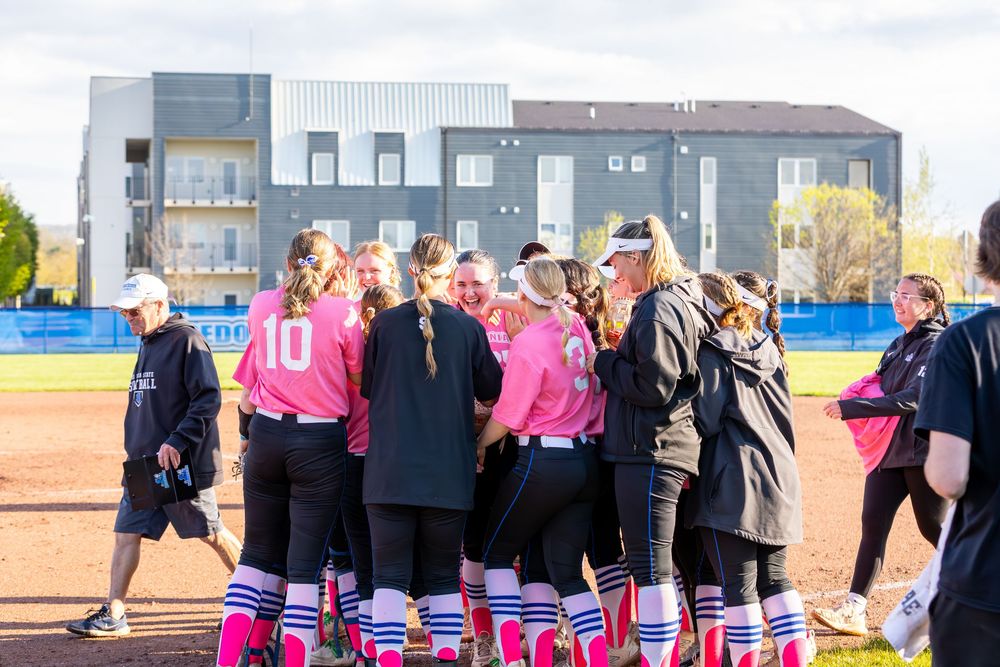 Teams in pink and black uniforms gather on softball field.
