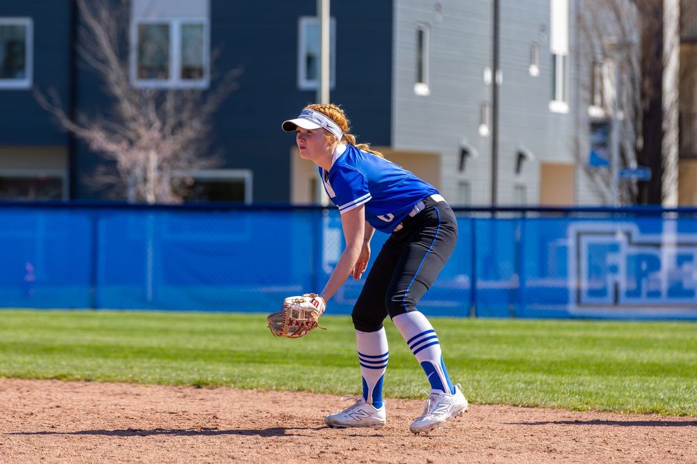 Softball players in action on field with buildings visible in background.