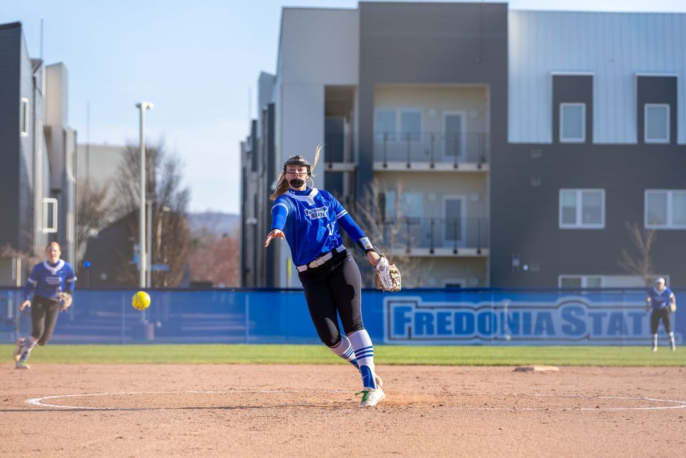 Softball play unfolding on dirt infield with yellow ball in motion.