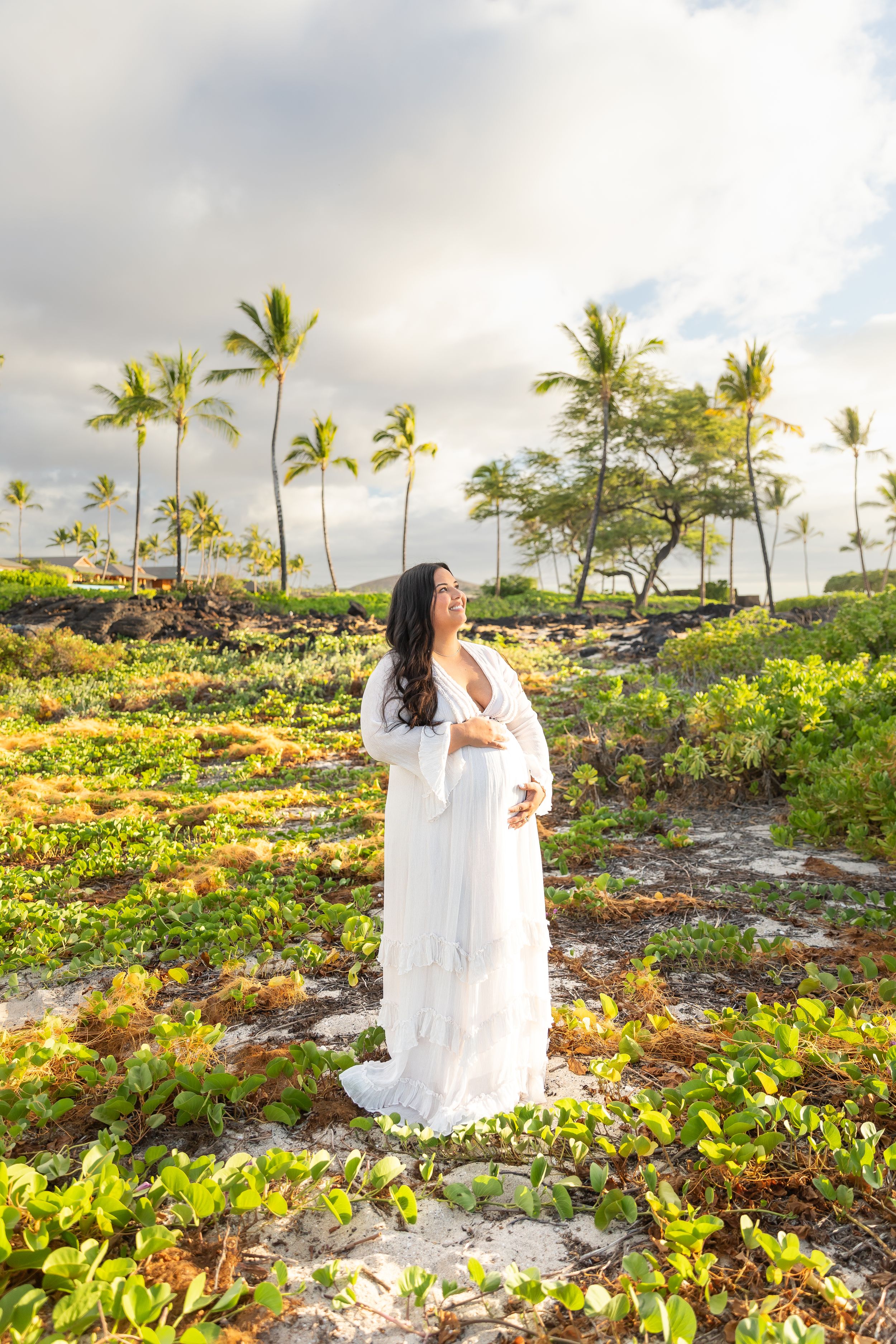 Ethereal maternity photo showing a white dress against swaying palm trees and lush tropical vegetation in Hawaii at sunset.