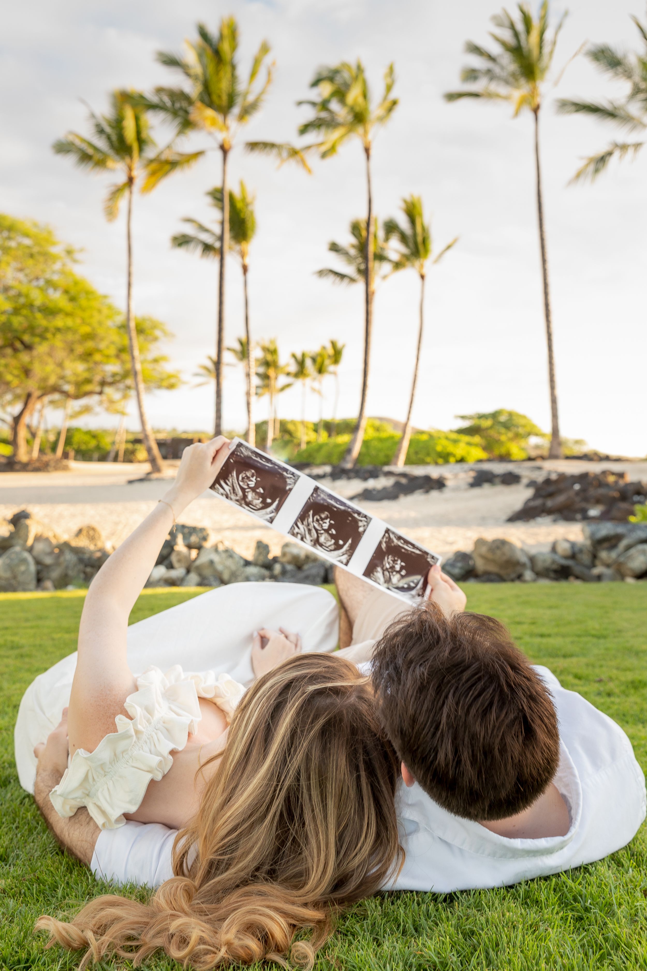 A couple lying on grass looking at sonogram photos with palm trees and ocean in the background at sunset.