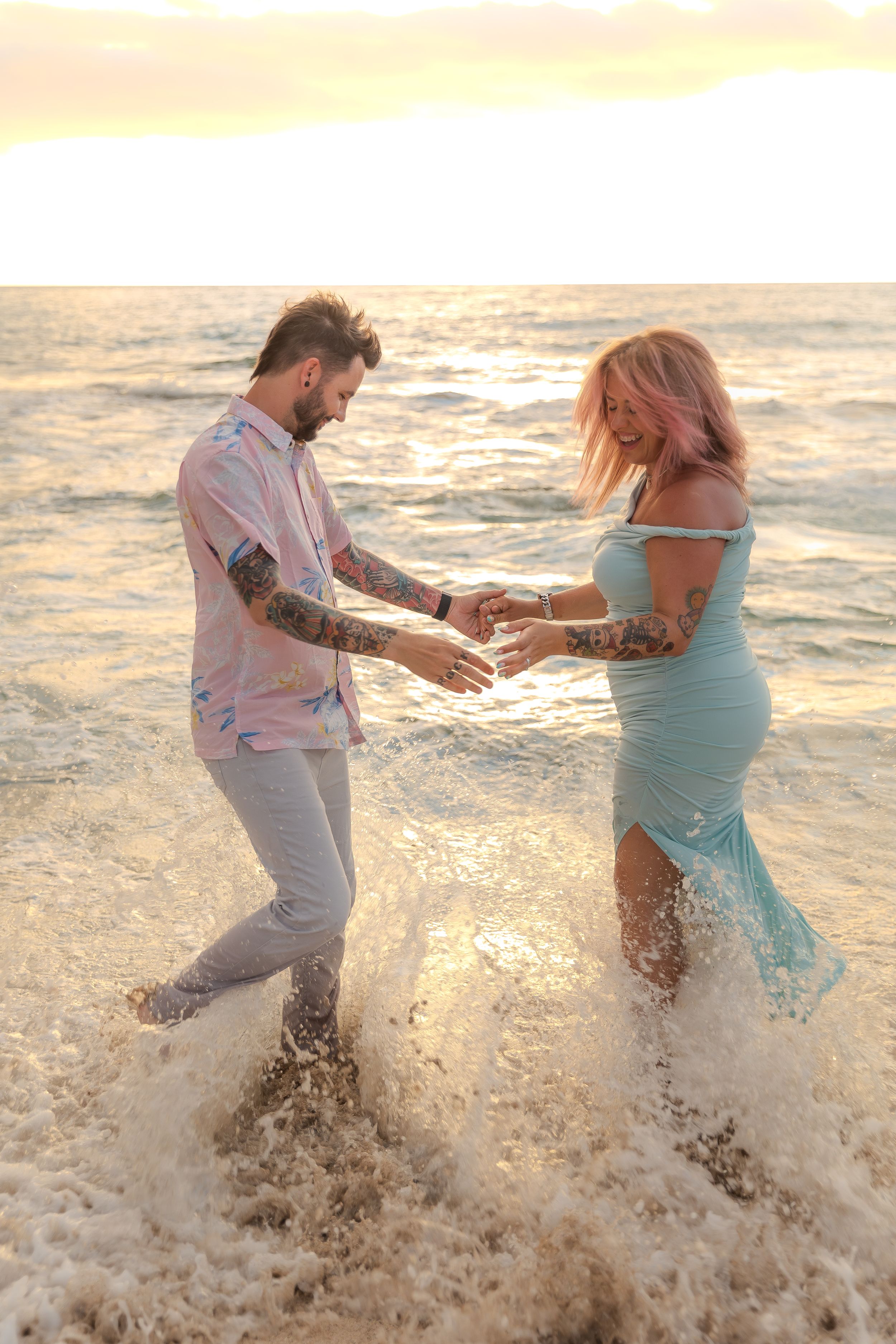 A couple splashes in the ocean waves during sunset, holding hands and dancing in turquoise and pink beach attire.