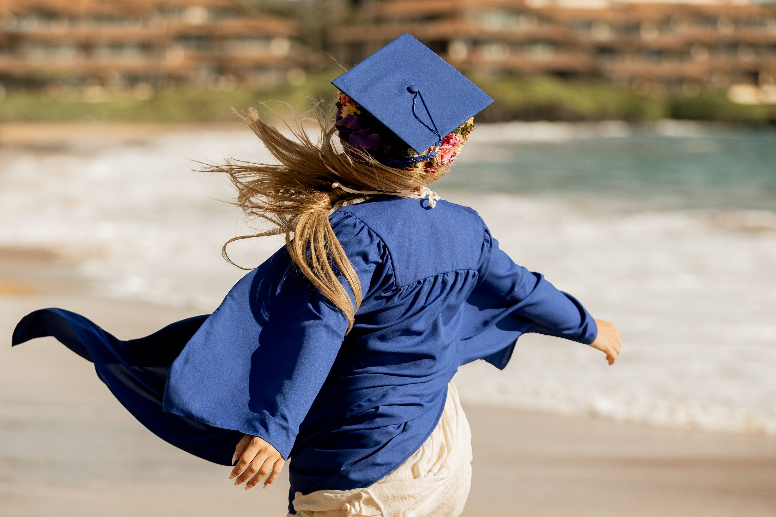 Person in blue graduation gown spins joyfully on beach.