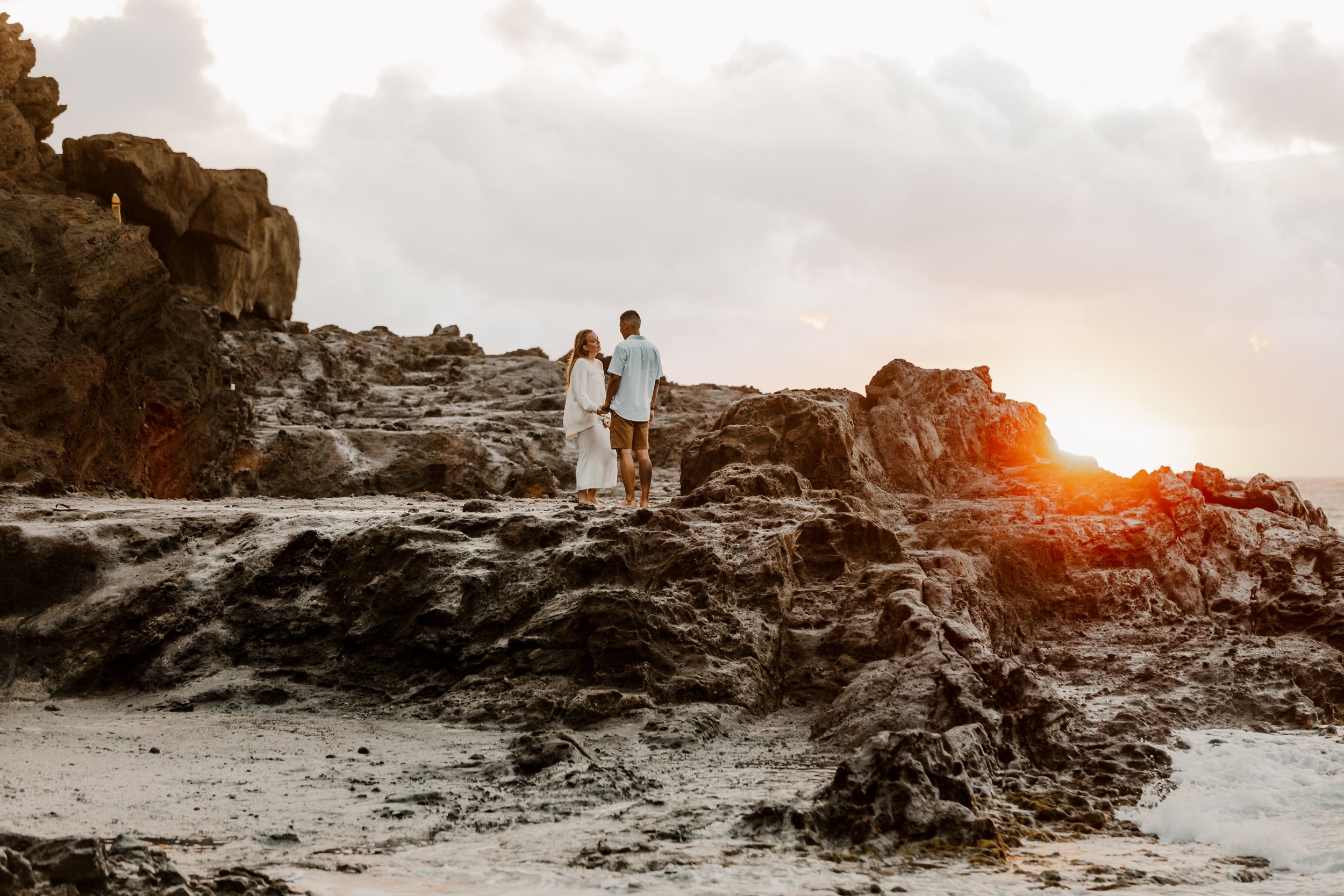 A couple stands on rocky cliffs at sunset overlooking the ocean.