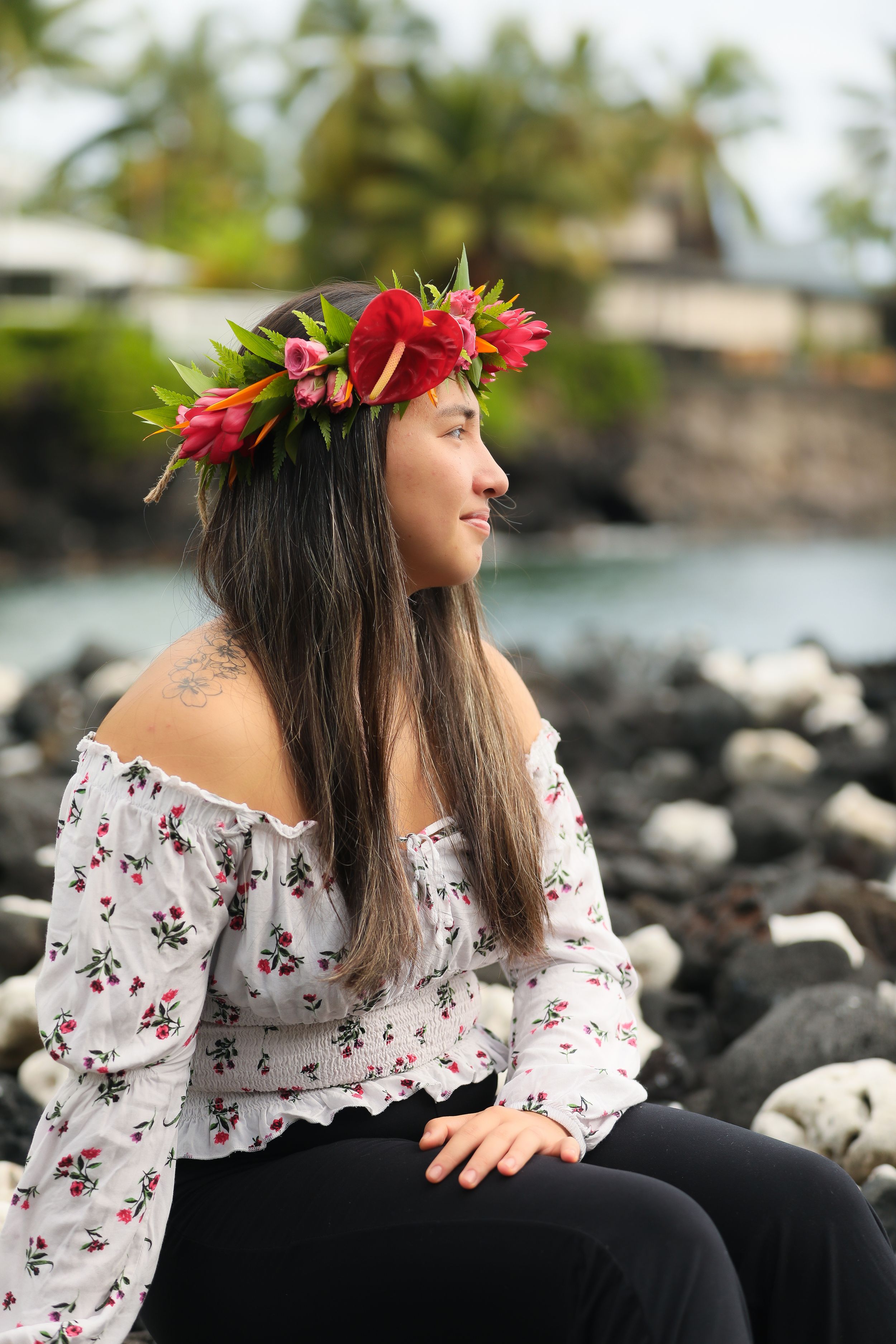 A woman in an off-shoulder floral top wearing a tropical flower crown sits on volcanic rocks by the ocean in Hawaii.