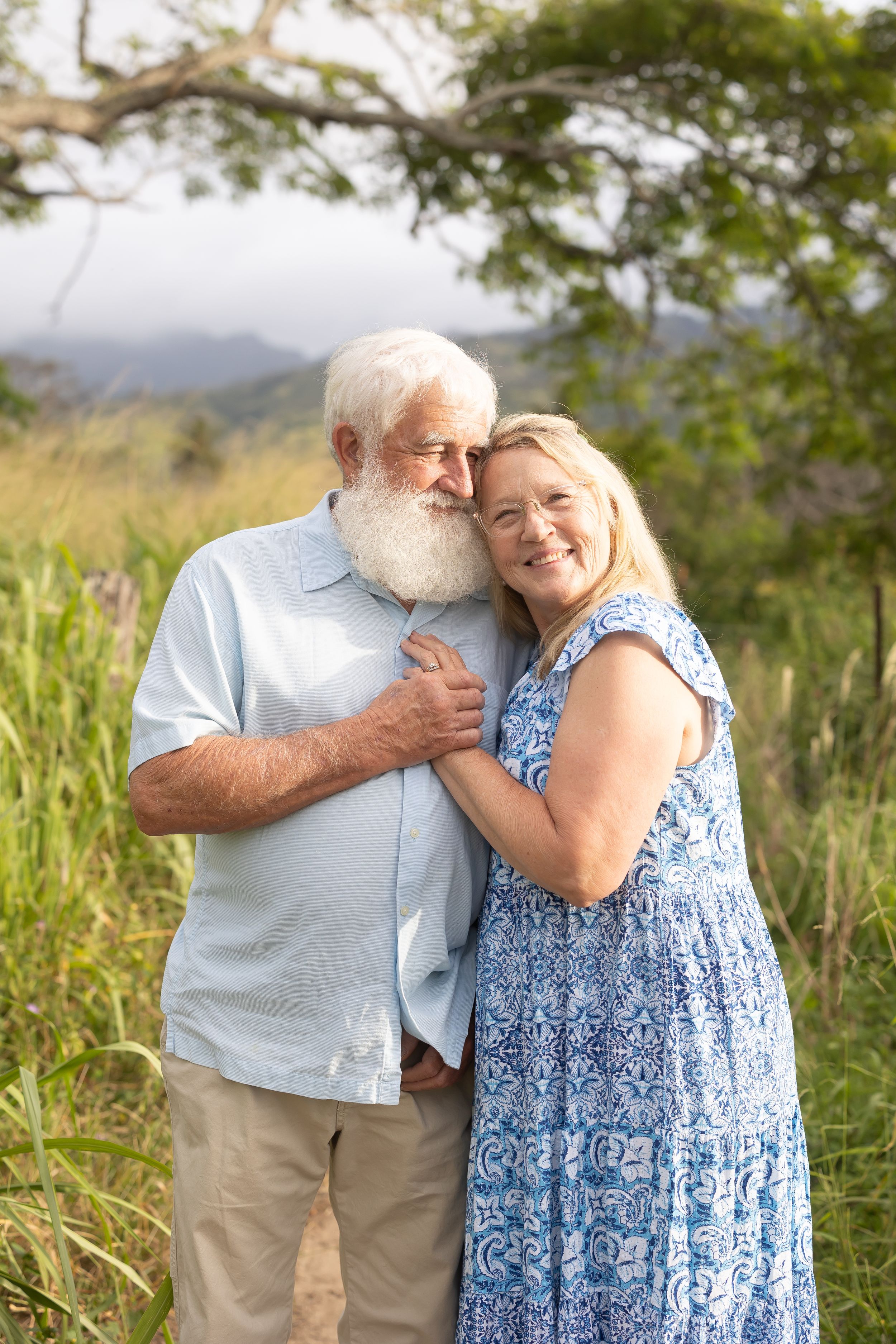 Elderly couple embracing outdoors on a scenic hillside with mountains and greenery in the background.