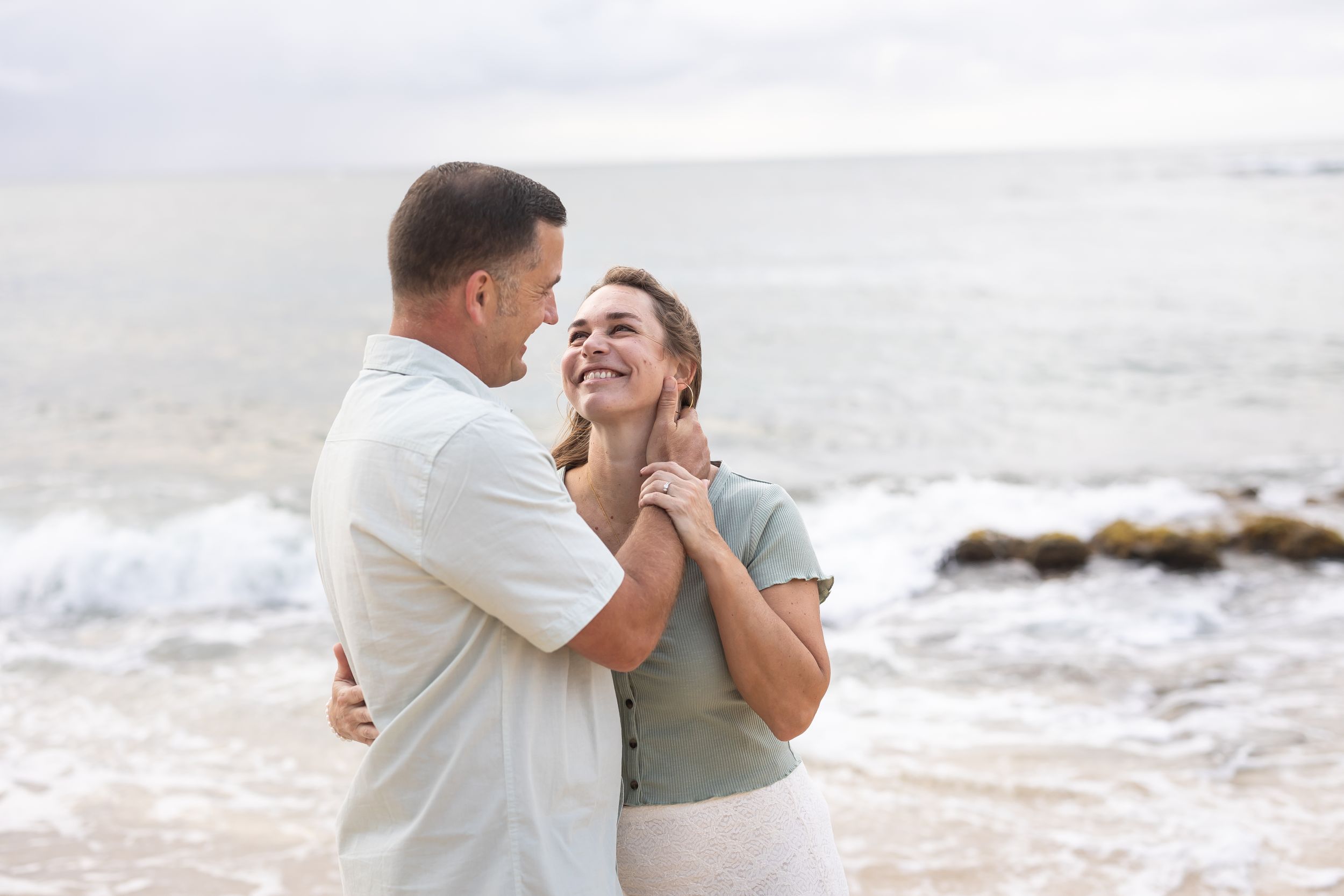 A romantic couple embraces and laughs together while standing on a misty beach at sunset.
