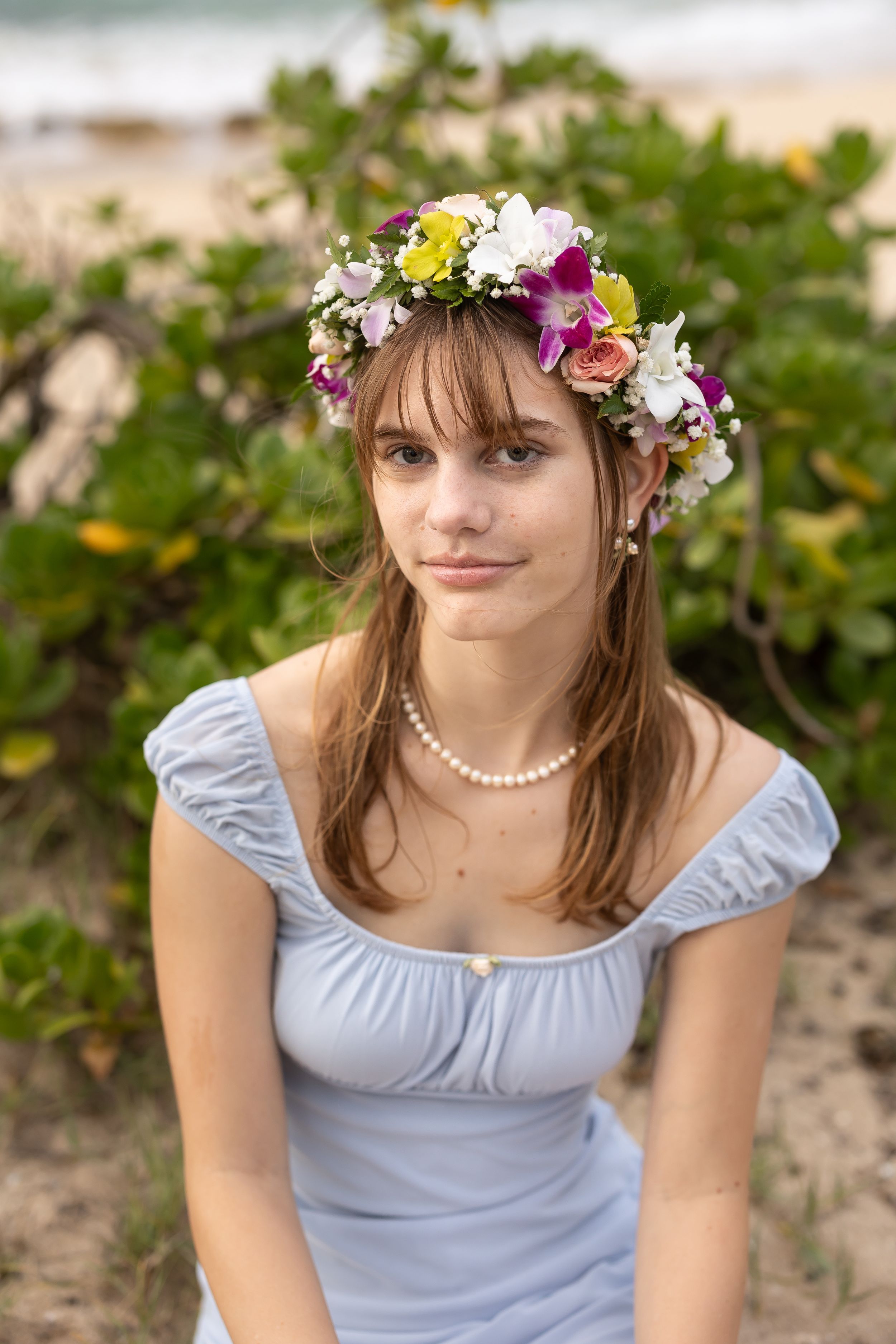 A floral crown adorned with colorful blooms sits atop brown hair, complementing a light blue summer dress with pearl necklace.