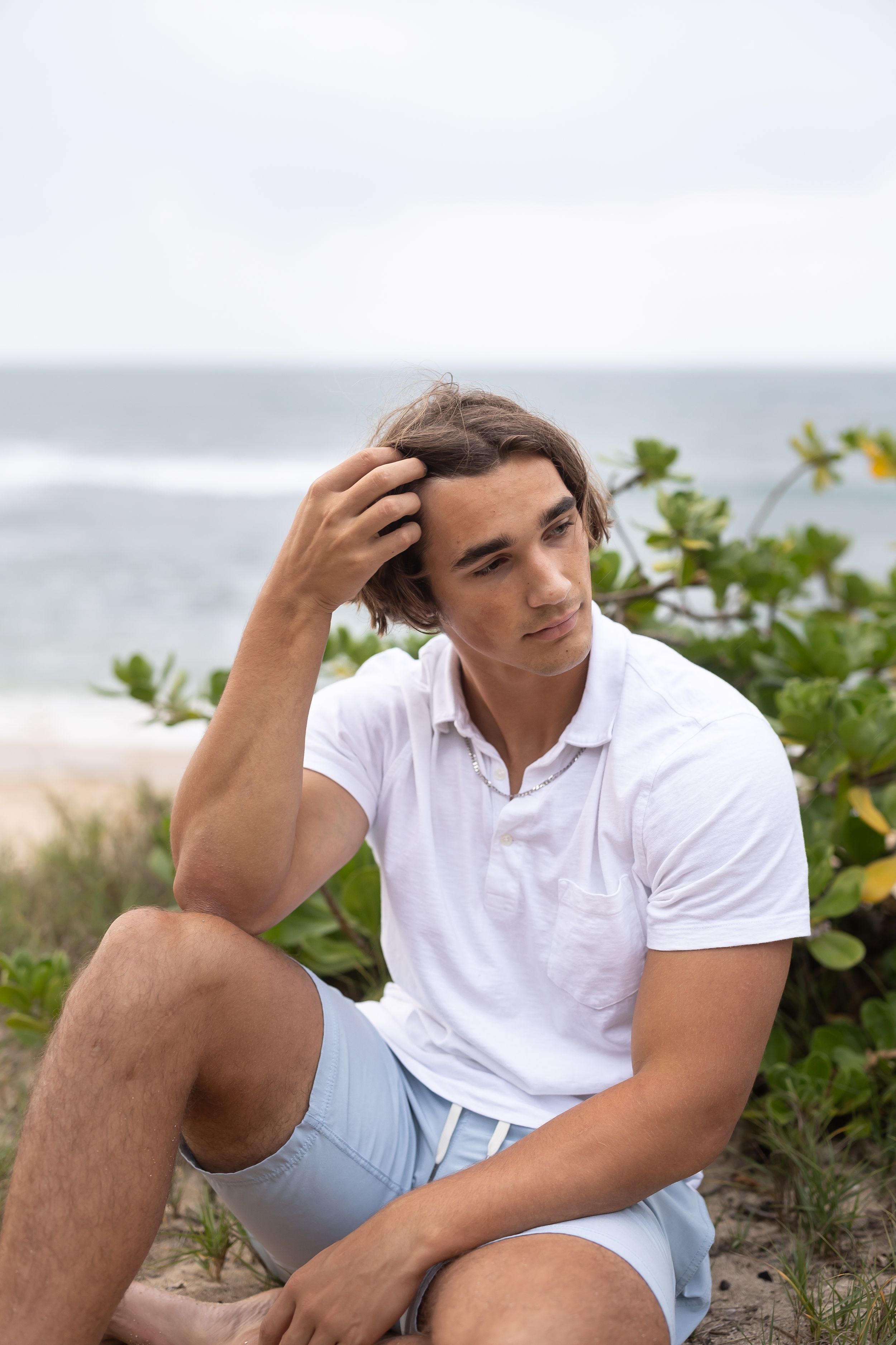 A person in a white polo shirt and shorts sits thoughtfully near the beach with foliage in the background.