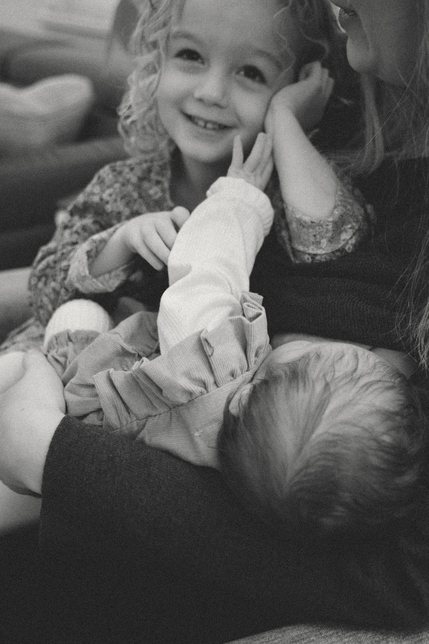 Black and white portrait of toddler girl looking to camera while cuddling her mom and her newborn sister during an at-home newborn session.