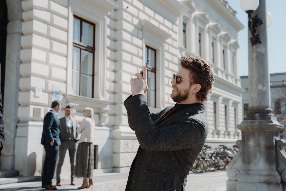 Veranstaltungsfotografie - Sponsion, Mann im Anzug hat ein Handy in der Hand und fotografiert lachend etwas.