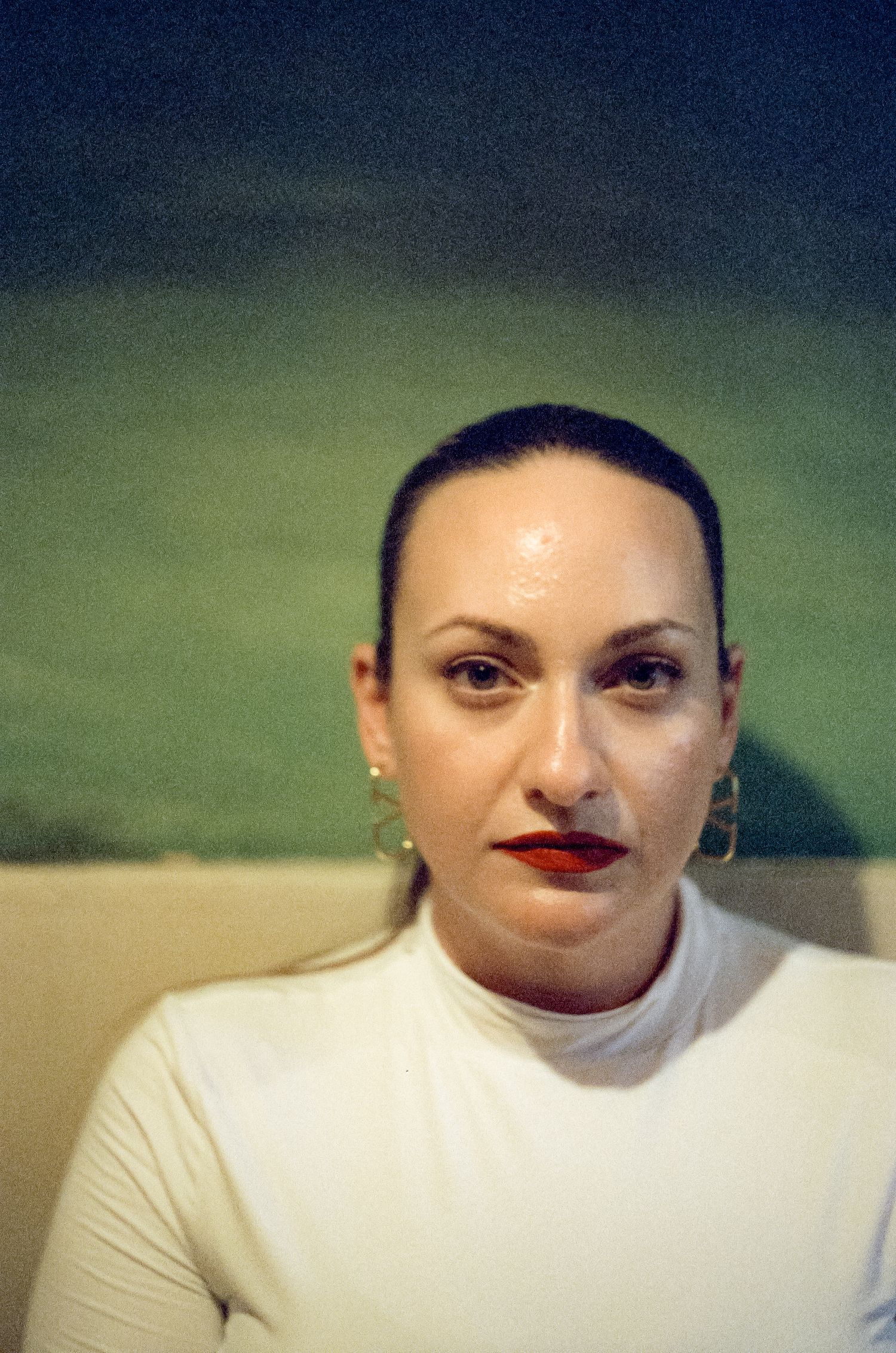 A close-up portrait shot of someone wearing a white top and red lipstick against a dark green background.