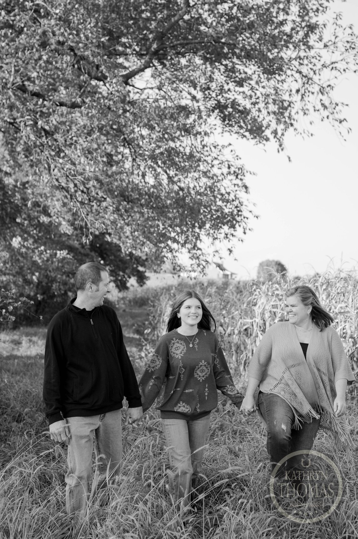 Three people walk together in tall grass beneath overhanging tree branches in black and white.