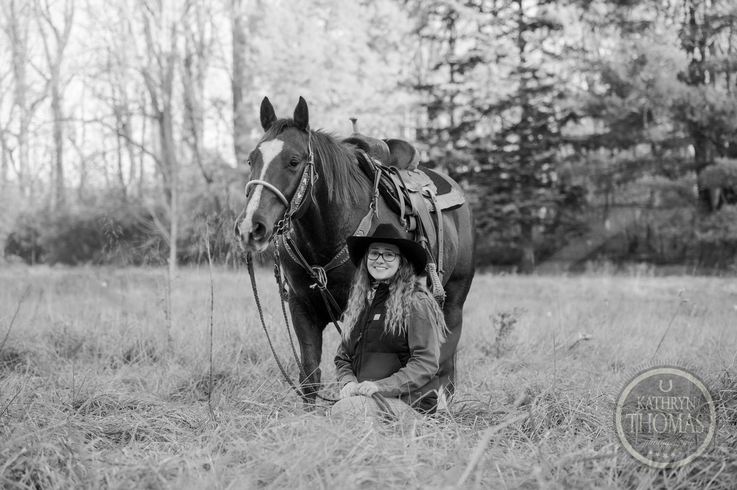 A black and white photo of a horse and rider in a field with evergreen trees in the background.