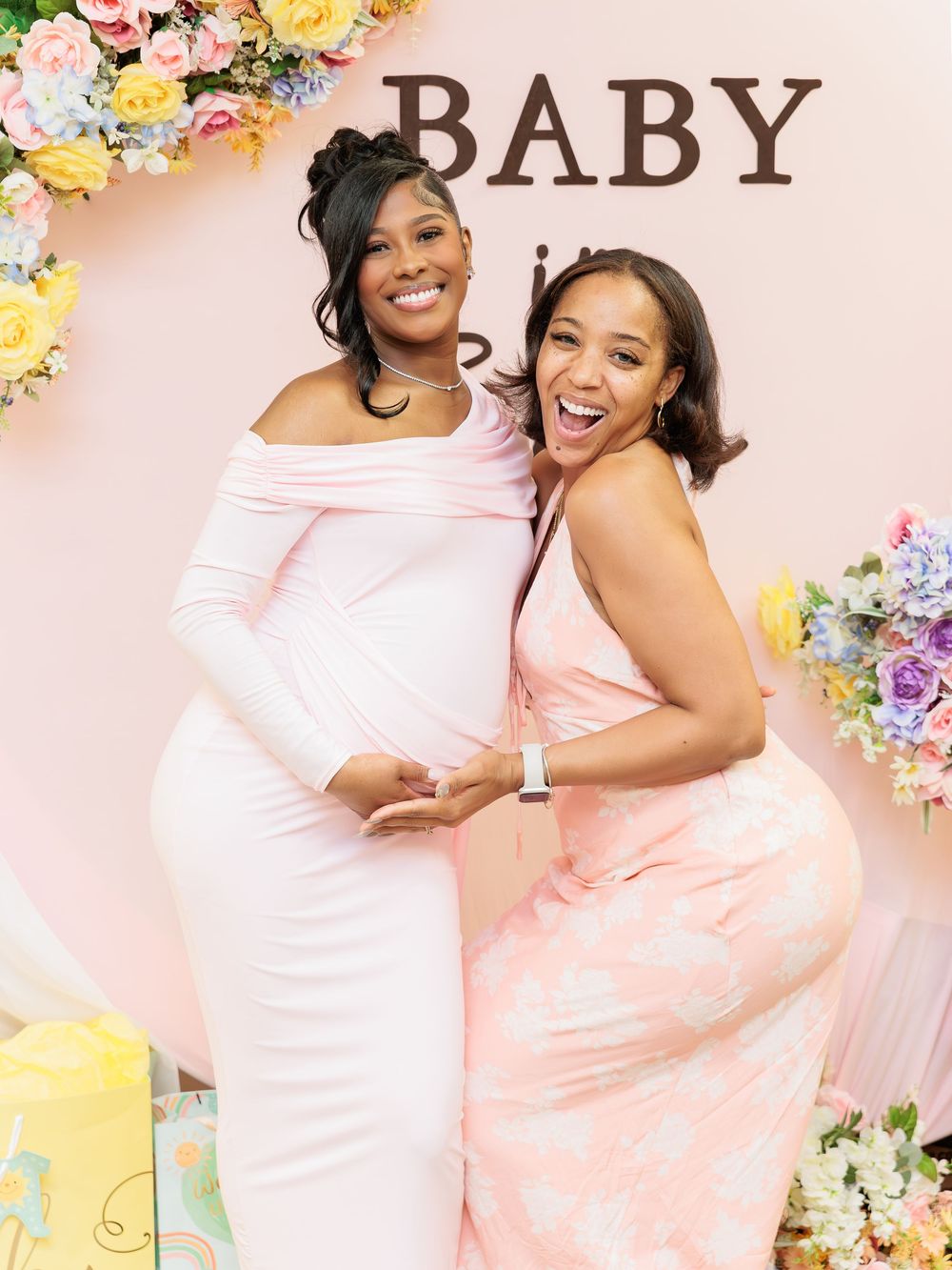 Two pregnant women in pink dresses smile and pose together at an elegant baby shower backdrop.
