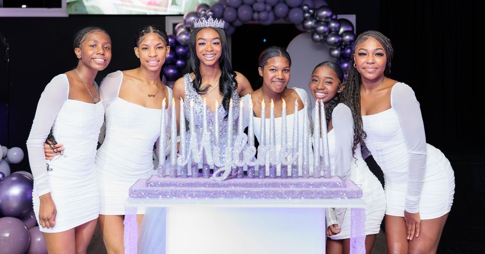 A birthday girl wearing a tiara poses with six friends in white dresses beside a decorated cake.