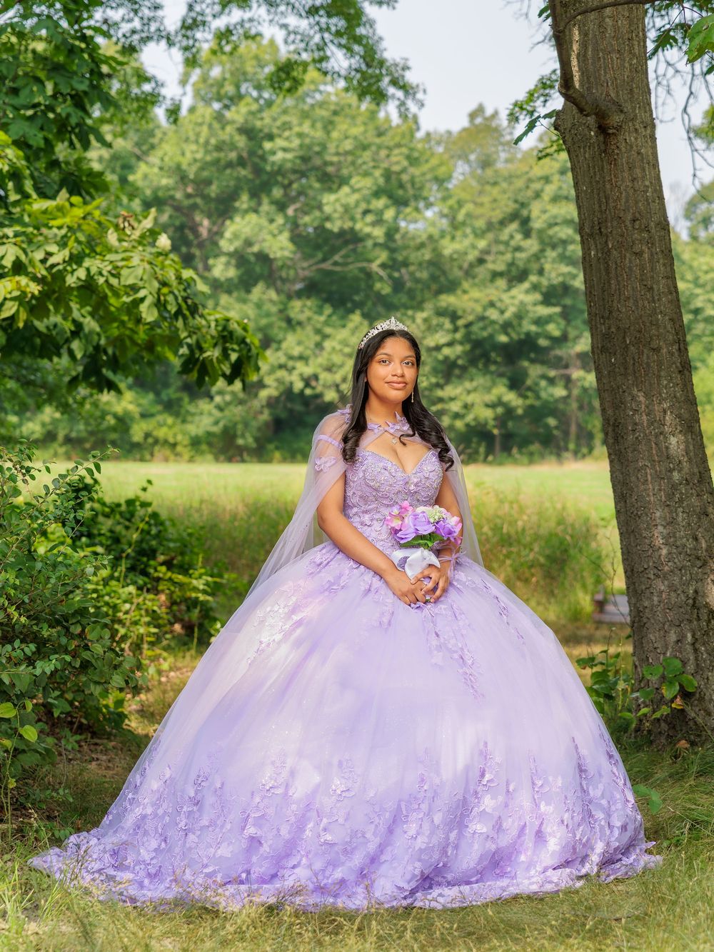 Young woman in lavender quinceañera ball gown holding bouquet in a lush green outdoor setting.