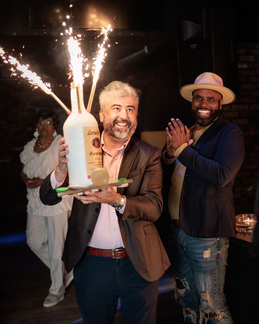 Man holding sparkler bottle cake at a celebration with guests applauding behind him.