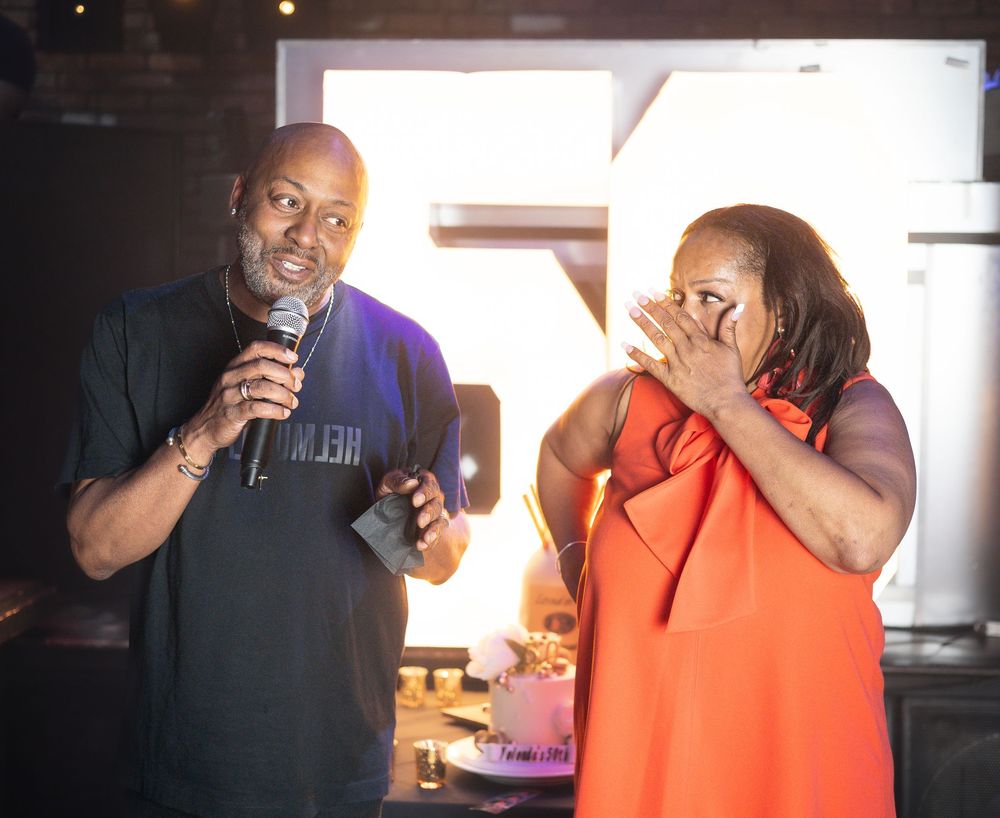 A man speaks into a microphone while a woman in an orange dress reacts emotionally beside him.