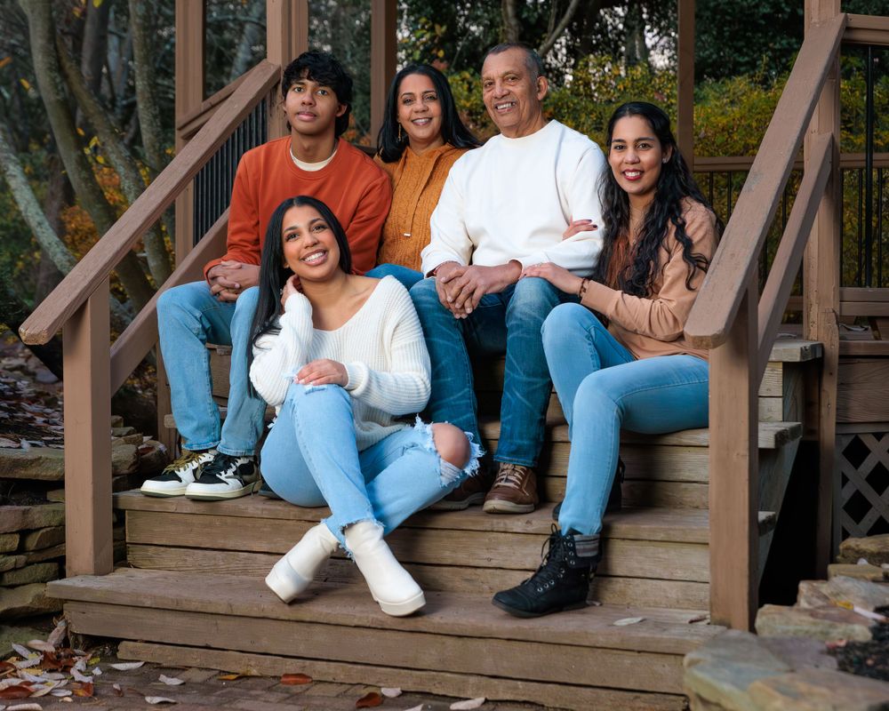 Smiling family of five sits closely together on wooden deck stairs in fall outdoor setting.
