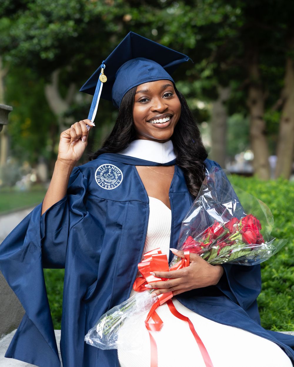 Smiling graduate in navy cap and gown holding roses and diploma tassel outdoors.