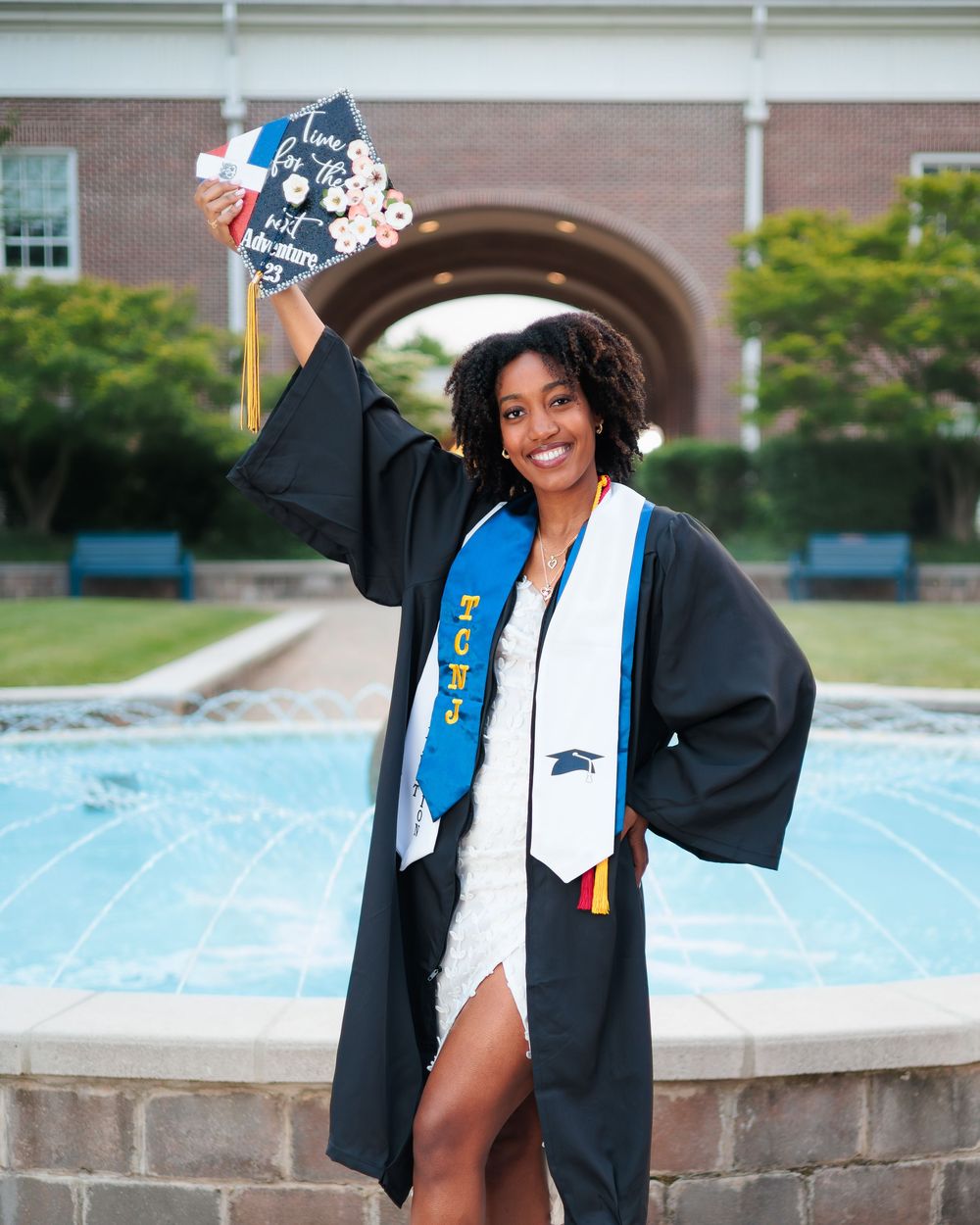 Graduate in black gown holding decorated cap beside a fountain at a college campus.