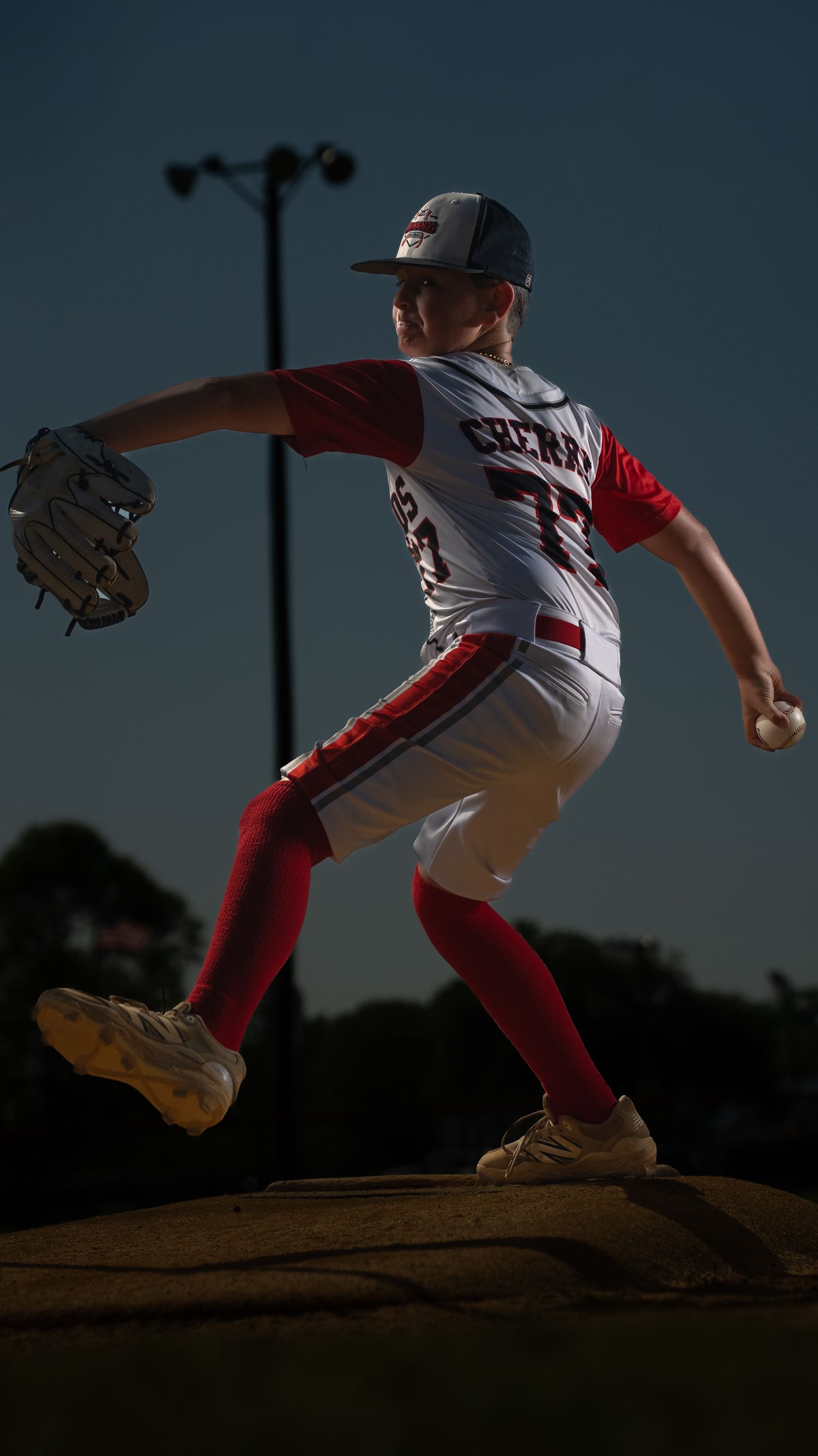 Young soccer player in white uniform stands with foot on pink ball against dramatic sunset sky.