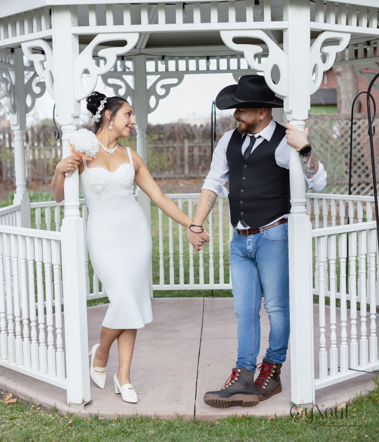 A couple poses romantically in a white gazebo, with the bride in a tea-length dress and the groom in western attire.