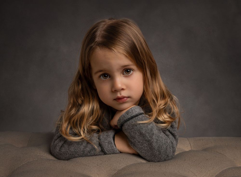 A small child smiling while leaning onto a bench in a photography studio with a soft grey background.