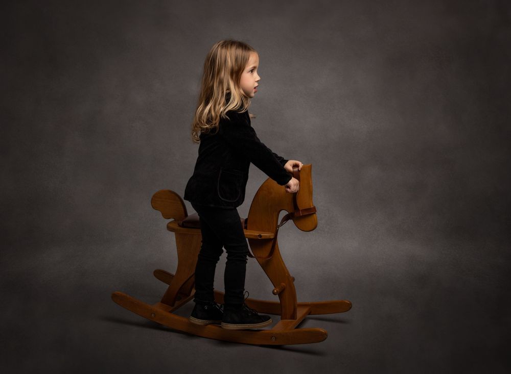 Fine-art studio portrait of a child on a vintage wooden rocking horse against a textured grey background.