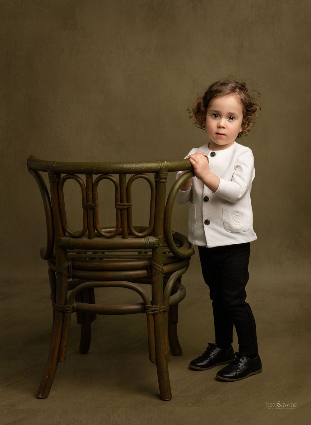 Fine art portrait of a boy standing next to a green wicker chair.