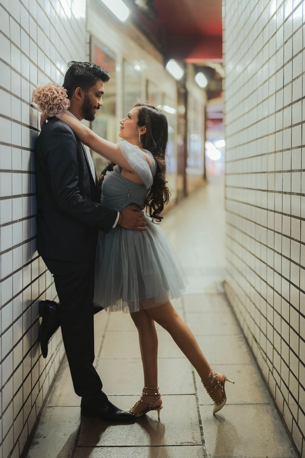 Couple leaning against tiled wall smiling at each other, woman in grey tulle dress holding flowers.
