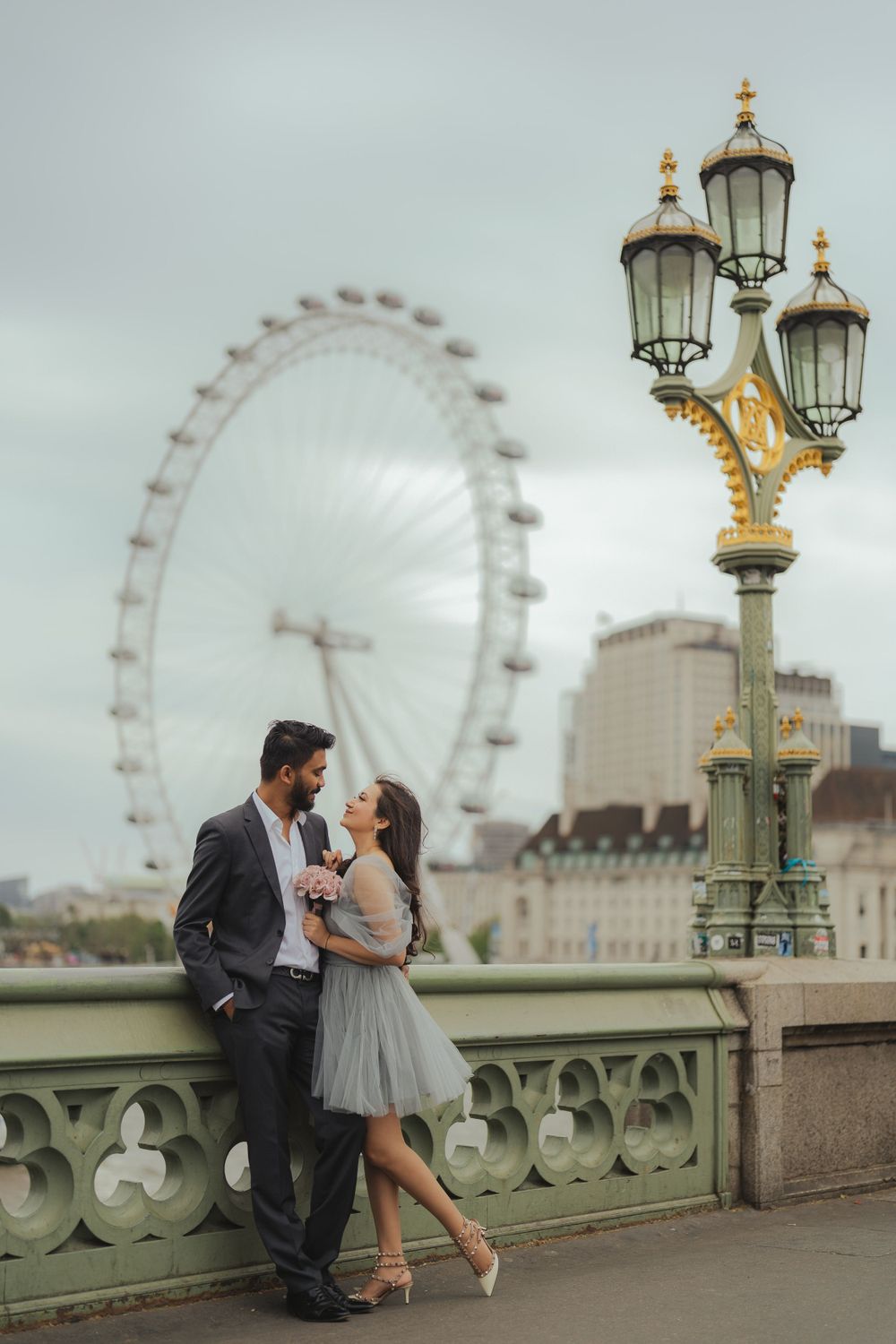 Couple posing near ornate lamppost on Westminster Bridge with London Eye in background.