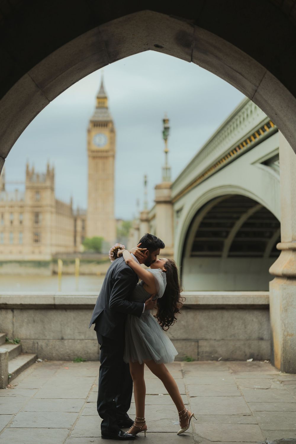 Couple sharing intimate kiss under arch with Big Ben and Westminster Bridge in background.