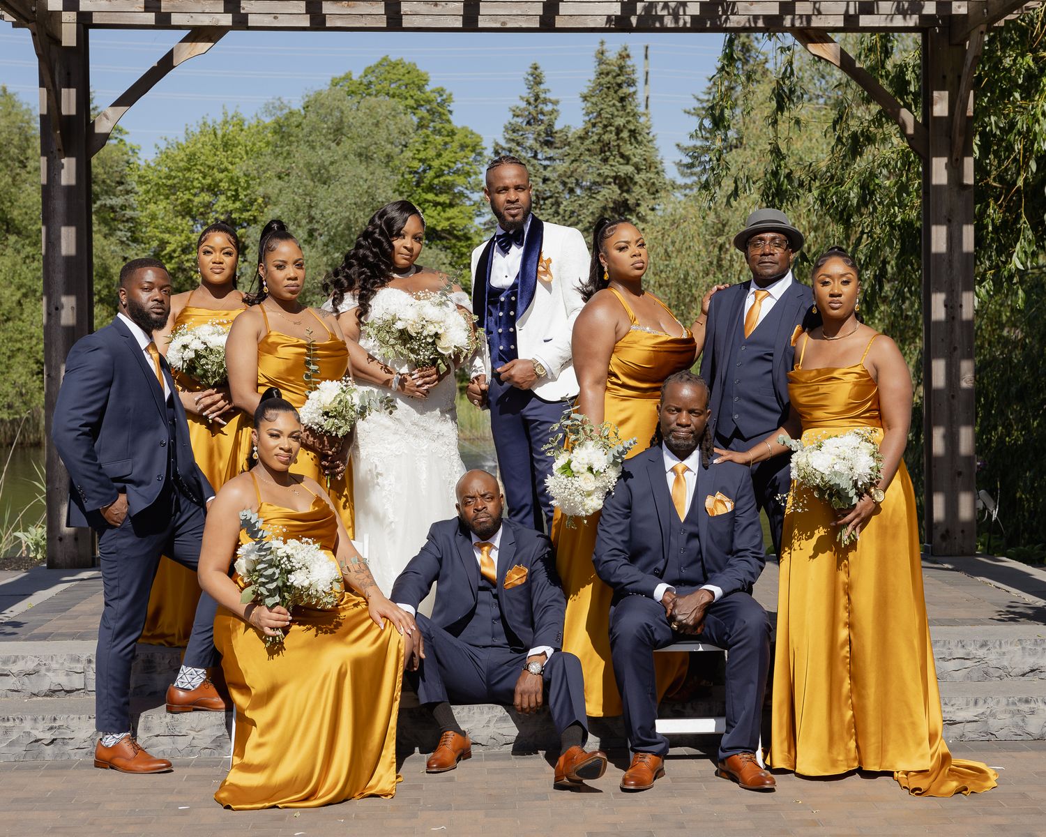 Wedding party group poses outdoors under wooden pergola wearing navy blue and mustard yellow attire with floral bouquets.