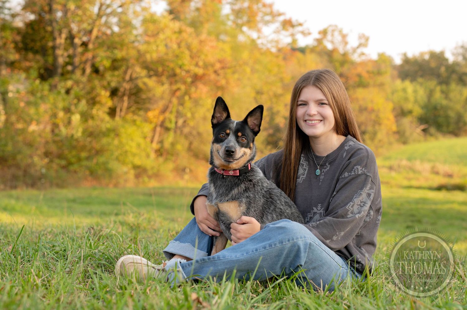 Person sits in grass with Australian Cattle Dog against autumn landscape backdrop.