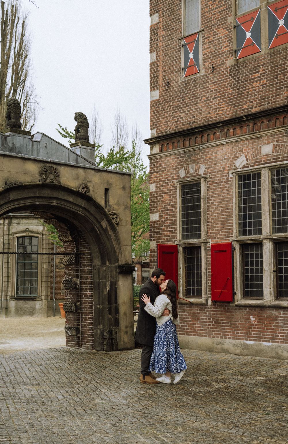 A couple embraces in front of a historic brick building with red shutters and arched doorways.